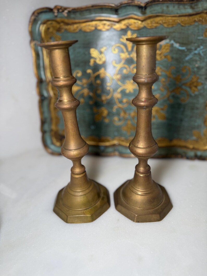 May include: Two antique brass candlesticks with a hexagonal base and a detailed, ornate design. The candlesticks have a warm, golden hue and are set against a blurred background.