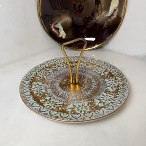 May include: A round metal serving tray with a central brass handle. The tray features a textured gold and light blue finish with a repeating heart and floral pattern along the rim. A dark brown, glossy, circular plate with gold edges is positioned behind the tray.