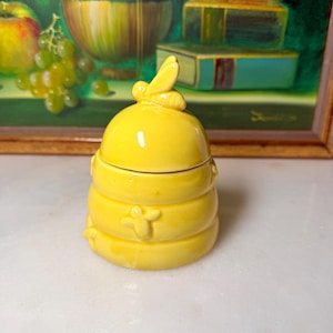 May include: A bright yellow ceramic honey pot shaped like a beehive, featuring a removable lid with a small sculpted bee on top and additional small bee figures embossed on the sides of the rounded, tiered body structure.