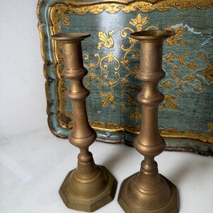 May include: Two antique brass candlesticks with a hexagonal base and ornate detailing. The candlesticks are a warm, golden color and stand in front of a teal and gold decorative tray.