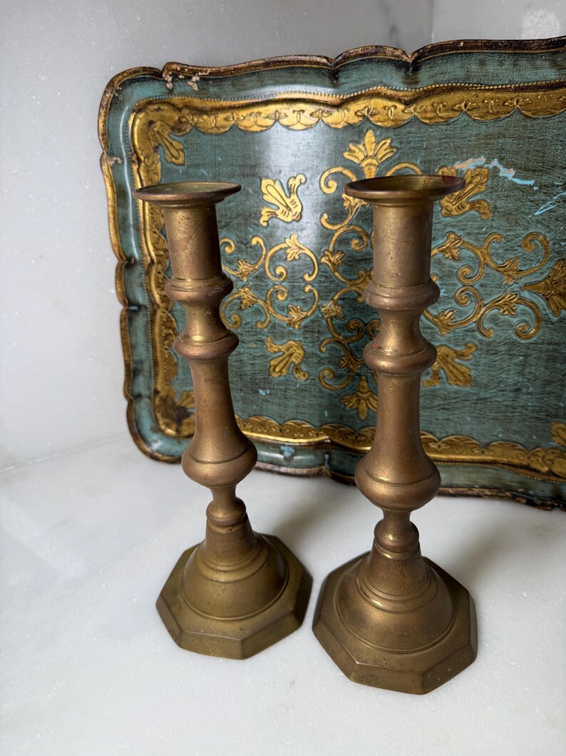 May include: Two antique brass candlesticks with a hexagonal base and ornate detailing. The candlesticks are a warm, golden color and stand in front of a teal and gold decorative tray.