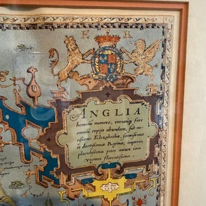 May include: Framed vintage map print of Anglia, featuring ornate text and illustrations. The map includes a coat of arms, lions, and text in Latin. The frame is a light brown color.