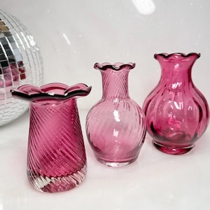 May include: Three small magenta glass vases with ruffled rims. The left vase has a diagonal ribbed pattern, the center vase features a slender neck with a subtle spiral texture, and the right vase has a rounded body with vertical ribbing.