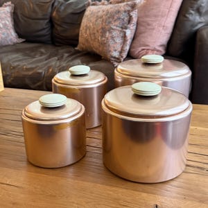May include: A set of three cylindrical copper-colored canisters with flat lids and small, pale green circular knobs on top. The canisters vary in size, with two smaller containers positioned behind a larger one, all featuring a smooth metallic finish.