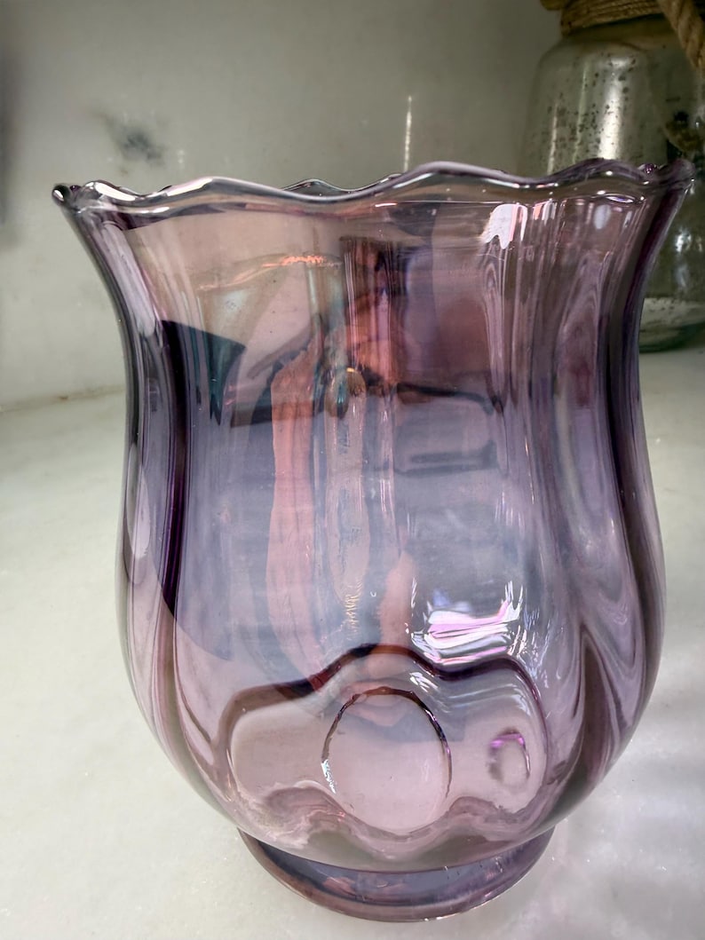 Peut inclure: Un vase d&eacute;coratif en verre violet iris&eacute; avec un bord festonn&eacute;. Le vase a un corps bulbeux et une ouverture &eacute;vas&eacute;e. Le verre pr&eacute;sente un motif tourbillonnant et une finition brillante, refl&eacute;tant la lumi&egrave;re et cr&eacute;ant un jeu de couleurs.