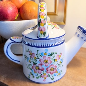 May include: White ceramic watering can with a floral design in pink, yellow, and blue. The handle and spout are accented with blue trim. A decorative piece for gardening or home decor.