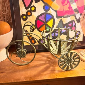 May include: A decorative metal tricycle planter in a distressed green finish. The planter features a basket for holding small plants or decorative items. The wheels and frame have intricate, stylized details. The background includes a colorful abstract painting.