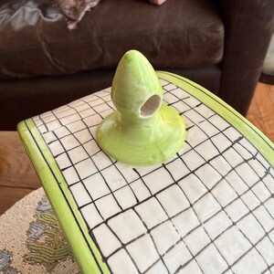 May include: A lime green ceramic decorative piece with a rounded top and a hole. The piece sits on a white surface with a black grid pattern and a green border. The background includes a brown leather couch and decorative pillows.