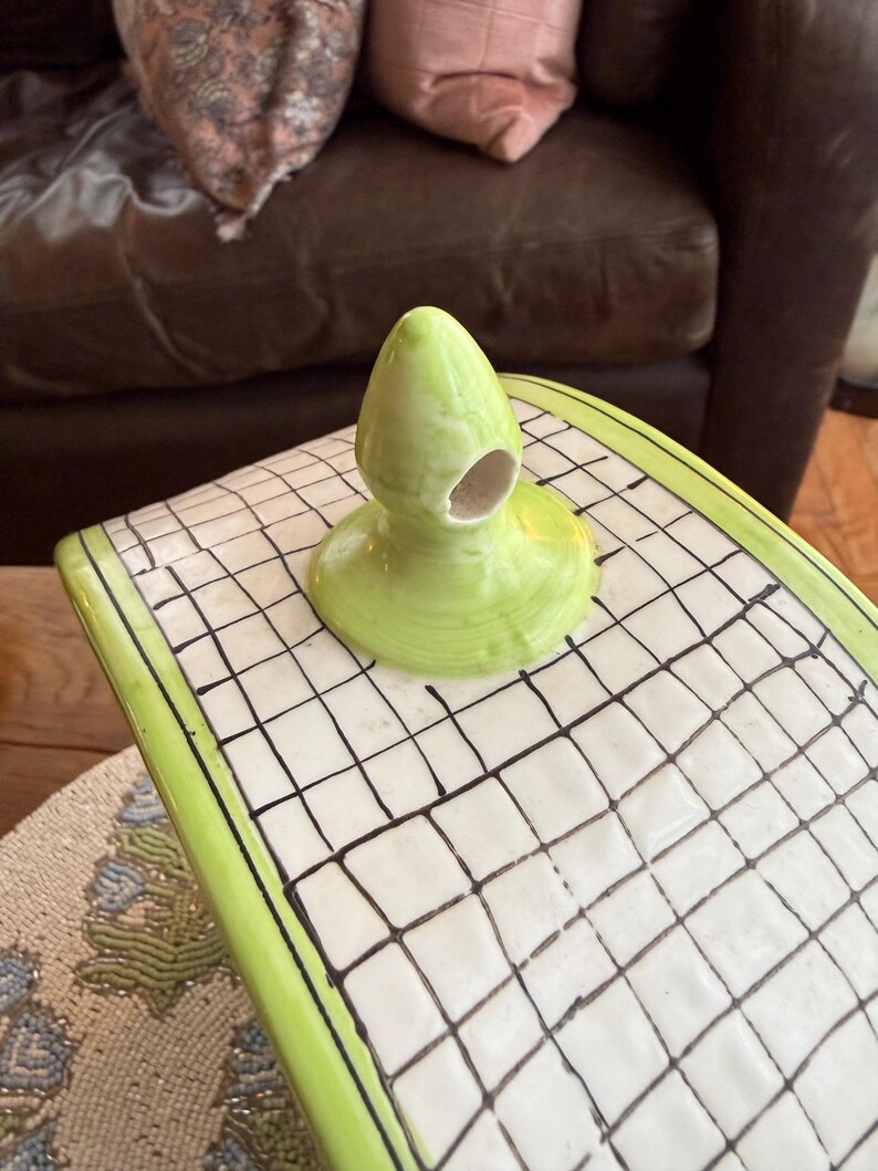 May include: A lime green ceramic decorative piece with a rounded top and a hole. The piece sits on a white surface with a black grid pattern and a green border. The background includes a brown leather couch and decorative pillows.