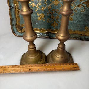 May include: Two antique brass candlesticks with a classic design. The candlesticks are approximately 10 inches tall. The base is a hexagonal shape. The background includes a decorative tray with gold and teal accents.