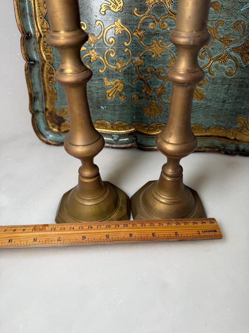 May include: Two antique brass candlesticks with a classic design. The candlesticks are approximately 10 inches tall. The base is a hexagonal shape. The background includes a decorative tray with gold and teal accents.