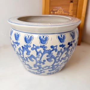 May include: A round ceramic planter featuring a cream-colored base with intricate blue floral and vine patterns. The rim is decorated with a geometric Greek key border. The bowl-shaped vessel has a wide opening and a smooth, glazed finish.