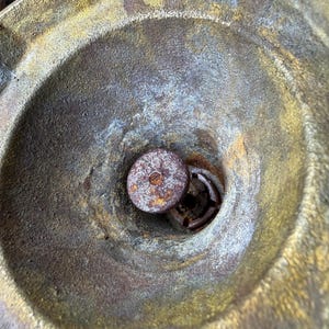 May include: Close-up of a weathered, antique metal object with a circular, bowl-like shape. The interior features a central, rusty metal component. The metal has a mix of gold, brown, and blue-grey tones, suggesting age and exposure.