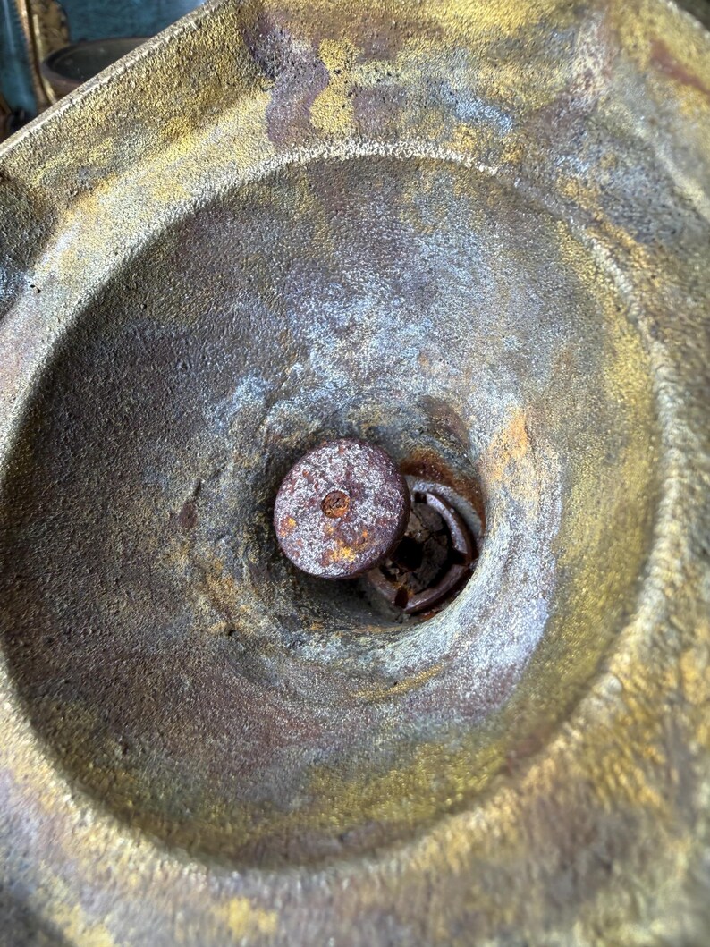 May include: Close-up of a weathered, antique metal object with a circular, bowl-like shape. The interior features a central, rusty metal component. The metal has a mix of gold, brown, and blue-grey tones, suggesting age and exposure.