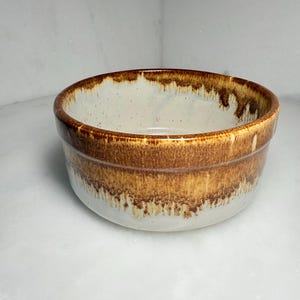 May include: A ceramic bowl with a white interior and a brown exterior. The bowl has a wide rim and a rounded shape. The brown color has a gradient effect, transitioning from a darker shade at the top to a lighter shade towards the bottom. The bowl is likely used for serving food.