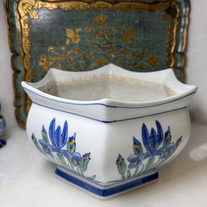 May include: White ceramic planter with a blue and white floral design. The planter has a square base and a scalloped rim. Blue iris flowers and green leaves decorate the sides. A blue and gold tray is in the background.