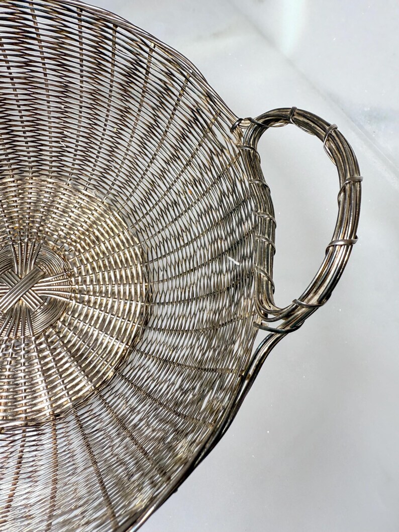 Puede incluir: Una cesta de servir plateada, tejida con alambre, con asa. La cesta tiene un dise&ntilde;o circular con un patr&oacute;n tejido que irradia desde el centro. El asa es un dise&ntilde;o curvo y en bucle, tambi&eacute;n hecho de alambre plateado.
