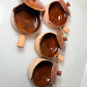 May include: Four small, round, ceramic cooking pots with lids. The pots are a light tan color on the outside and a glossy brown on the inside. Each pot has a handle and a matching lid with a small knob.
