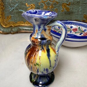 May include: A small, hand-painted ceramic pitcher with a handle. The pitcher features a bulbous body and a flared spout, with a base. The colors include blue, yellow, brown, and green, creating a unique, artistic appearance. The pitcher is a decorative item.