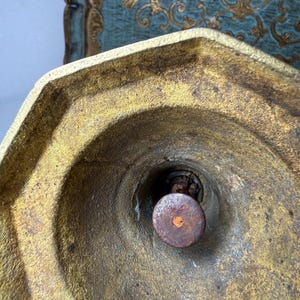 May include: Close-up of a gold-colored, octagonal metal object with a central, circular, rusty brown component. The object has a weathered appearance, with visible wear and tear. The background features a teal surface with gold decorative elements.