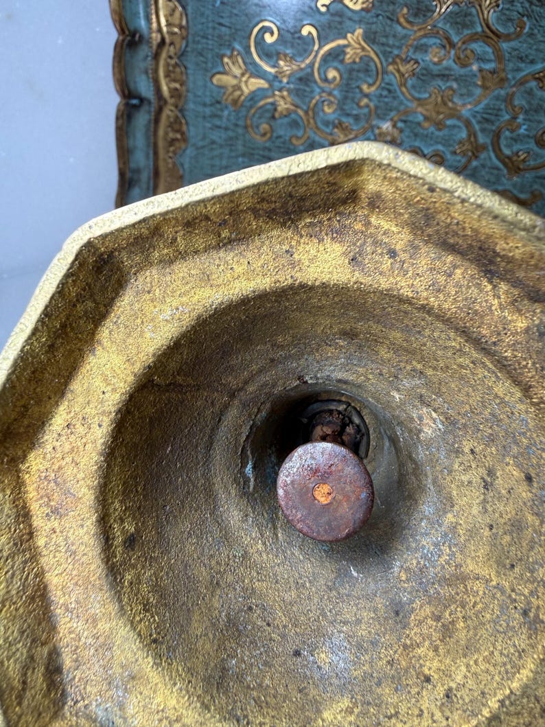 May include: Close-up of a gold-colored, octagonal metal object with a central, circular, rusty brown component. The object has a weathered appearance, with visible wear and tear. The background features a teal surface with gold decorative elements.