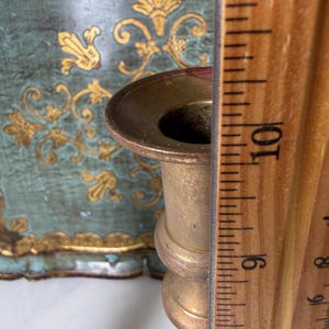 May include: A brass candlestick with a round top and a decorative base. A wooden ruler is placed next to the candlestick, showing a measurement of approximately 9 inches. The background features a teal and gold decorative panel.