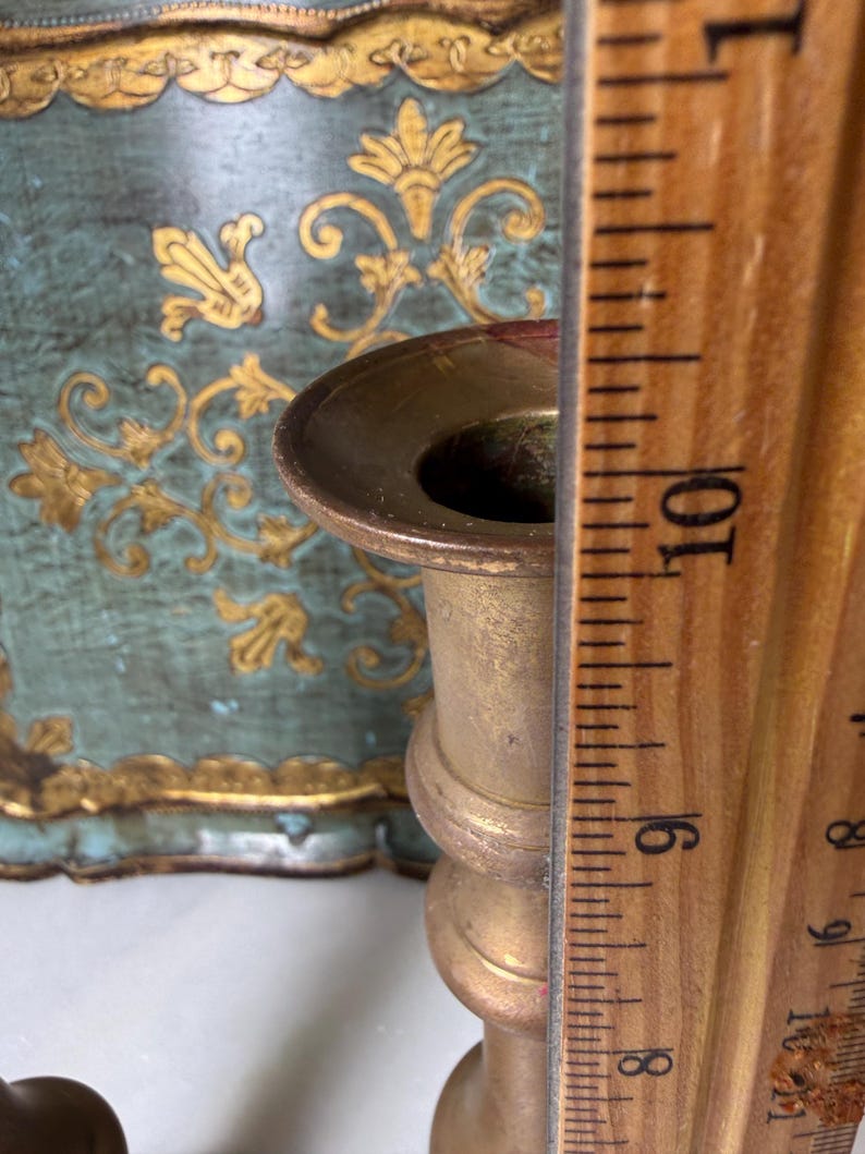 May include: A brass candlestick with a round top and a decorative base. A wooden ruler is placed next to the candlestick, showing a measurement of approximately 9 inches. The background features a teal and gold decorative panel.