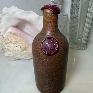 May include: A brown ceramic bottle with a dark red wax seal and stopper. The bottle has a textured surface and a decorative seal near the base. A clear glass is visible in the background.
