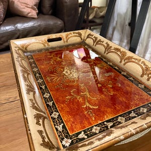May include: A rectangular serving tray with a glossy finish, featuring a central orange and brown marbled panel decorated with gold floral motifs. The cream-colored border is adorned with intricate gold scrollwork and black accents.