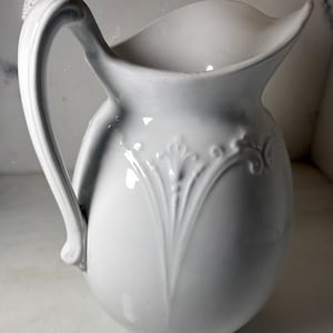 Può includere: Una brocca in ceramica bianca con un manico curvo e un design decorativo in rilievo. La brocca ha un beccuccio largo e una finitura liscia e lucida. Il design in rilievo è un motivo floreale stilizzato.