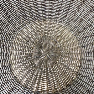 Puede incluir: Primer plano de una cesta de metal plateado con un dise&ntilde;o tejido. El patr&oacute;n de la cesta irradia desde el centro, creando un efecto de espiral. El metal tiene una calidad brillante y reflectante, y el tejido es intrincado.