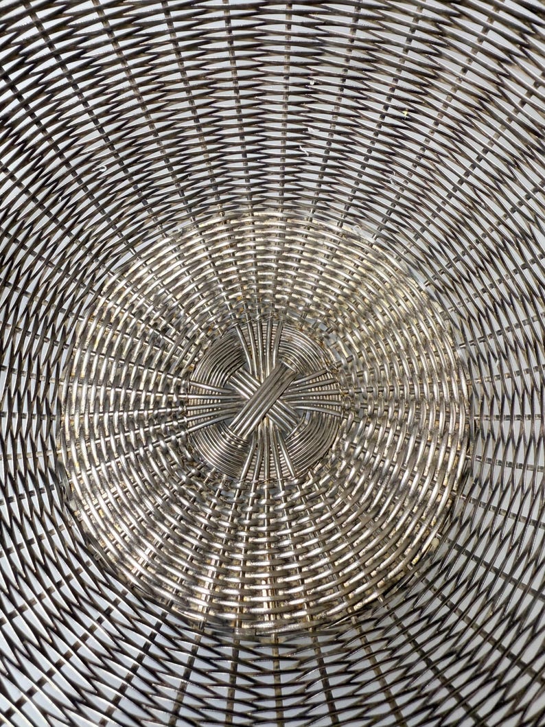 Puede incluir: Primer plano de una cesta de metal plateado con un dise&ntilde;o tejido. El patr&oacute;n de la cesta irradia desde el centro, creando un efecto de espiral. El metal tiene una calidad brillante y reflectante, y el tejido es intrincado.
