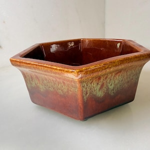 May include: A square, ceramic planter with a rich, reddish-brown glaze. The planter has a slightly flared rim and a band of green and brown hues near the base. The interior is a darker shade of brown.