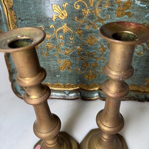 May include: Two antique brass candlesticks with a classic design. The candlesticks have a wide base and a flared top. The background features a teal tray with gold floral accents. The candlesticks are a warm gold color.