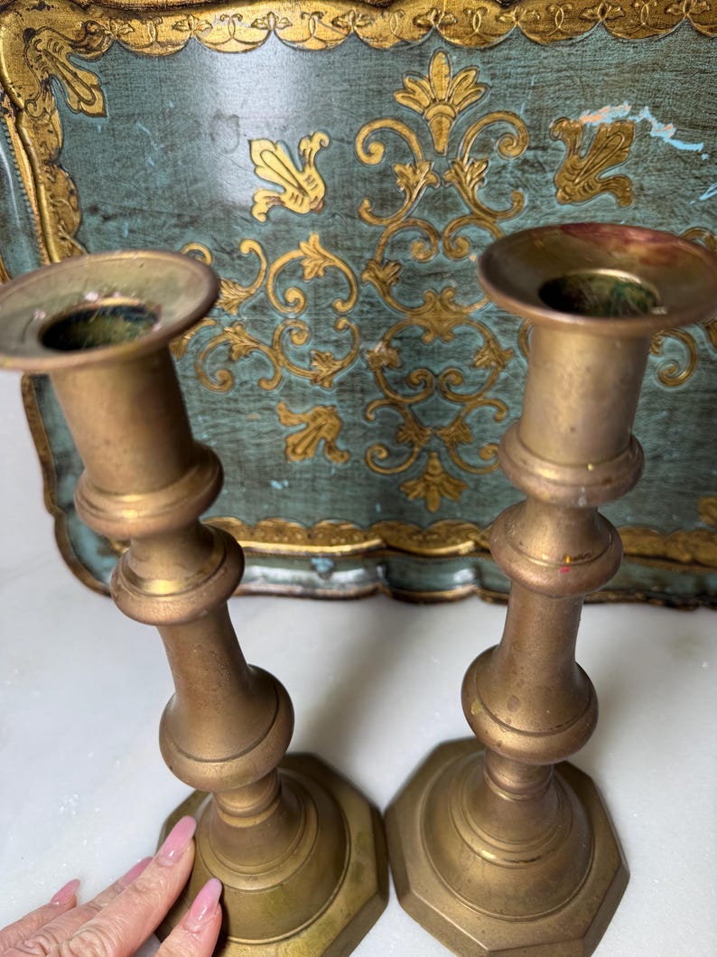 May include: Two antique brass candlesticks with a classic design. The candlesticks have a wide base and a flared top. The background features a teal tray with gold floral accents. The candlesticks are a warm gold color.