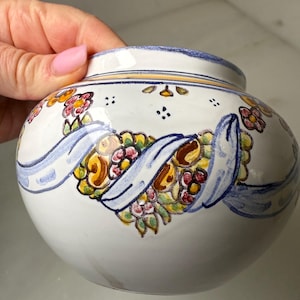 May include: White ceramic vase with a rounded shape, adorned with floral and ribbon designs in shades of yellow, pink, and blue. The vase has a blue rim and is held by a hand with pink nail polish.