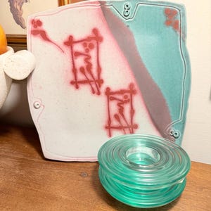 May include: A square ceramic plate with a white and pink surface, featuring red abstract designs and a turquoise border. A stack of clear, teal-colored glass rings sits in front of the plate.