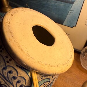 May include: A ceramic lamp base with a cream-colored top and a large central opening. The base is decorated with blue and white floral patterns. The lamp has a cord and is partially visible.