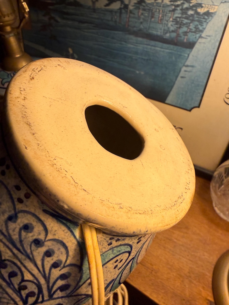 May include: A ceramic lamp base with a cream-colored top and a large central opening. The base is decorated with blue and white floral patterns. The lamp has a cord and is partially visible.