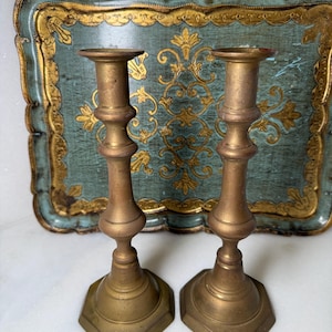 May include: Two antique brass candlesticks with a hexagonal base and ornate detailing. The candlesticks are displayed in front of a vintage teal tray with gold accents. The tray has a scalloped edge and a decorative floral pattern.