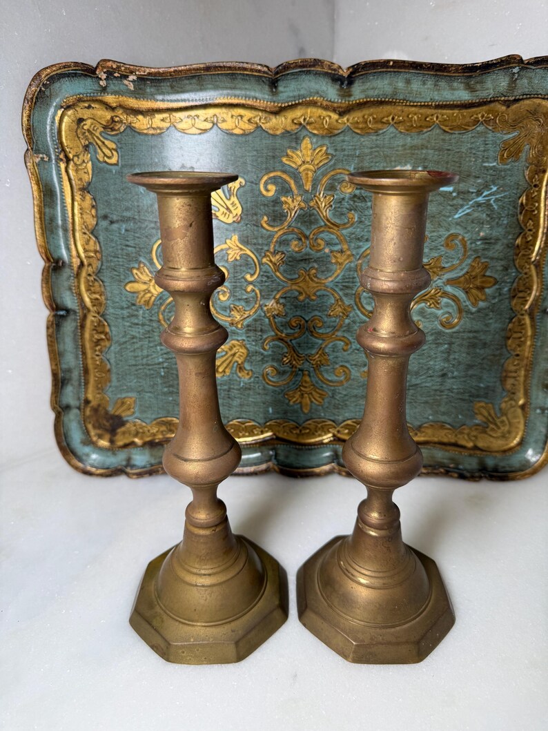 May include: Two antique brass candlesticks with a hexagonal base and ornate detailing. The candlesticks are displayed in front of a vintage teal tray with gold accents. The tray has a scalloped edge and a decorative floral pattern.