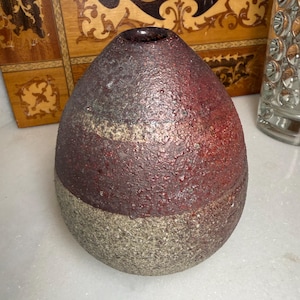 May include: A rounded ceramic vase with a narrow opening at the top. The upper portion features a textured, deep reddish-brown glaze, while the lower portion has a coarse, speckled grey stone-like finish. The surface has a rough, tactile quality.