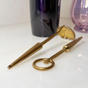Peut inclure: Deux outils de bar vintage en laiton : une cuill&egrave;re &agrave; fentes et un ouvre-bouteille. La cuill&egrave;re a une t&ecirc;te ronde et perfor&eacute;e et un long manche. L'ouvre-bouteille est dot&eacute; d'une poign&eacute;e en anneau. Les deux outils ont une teinte dor&eacute;e et sont pos&eacute;s sur un fond blanc.