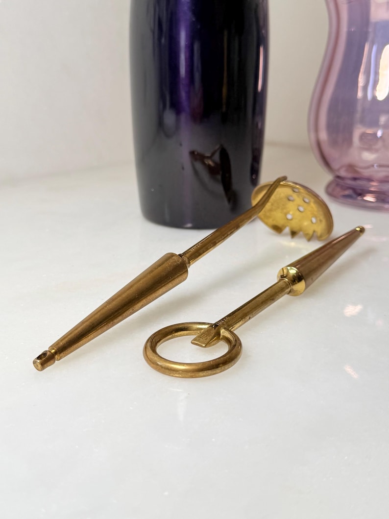 Peut inclure: Deux outils de bar vintage en laiton : une cuill&egrave;re &agrave; fentes et un ouvre-bouteille. La cuill&egrave;re a une t&ecirc;te ronde et perfor&eacute;e et un long manche. L'ouvre-bouteille est dot&eacute; d'une poign&eacute;e en anneau. Les deux outils ont une teinte dor&eacute;e et sont pos&eacute;s sur un fond blanc.