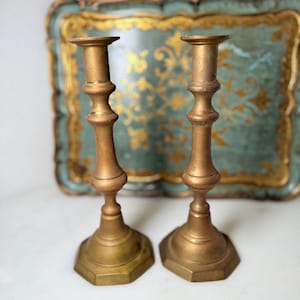 May include: Two antique brass candlesticks. Each candlestick has a hexagonal base, a slender, ornate stem, and a circular top for holding a candle. The candlesticks are a warm, aged brass color, suggesting they are vintage or antique.