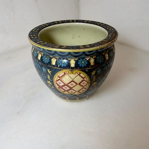 May include: A small, round ceramic planter featuring a dark blue base with intricate patterns of light blue flowers, yellow accents, and a central cream-colored medallion with a red and white diamond grid design and a pale green interior.