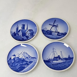 May include: Set of four blue and white ceramic coasters. Each coaster features a different scenic design: a church, a windmill, mountains, and a ship. The coasters have a white border and are round.