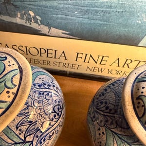May include: Two ceramic lamps with blue and white floral patterns. The lamps have a round base and a matching top. The background includes a framed print with the text "CASSIOPEIA FINE ART" and "Bleecker Street New York".