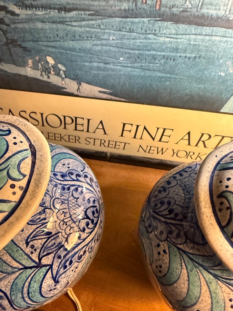 May include: Two ceramic lamps with blue and white floral patterns. The lamps have a round base and a matching top. The background includes a framed print with the text "CASSIOPEIA FINE ART" and "Bleecker Street New York".