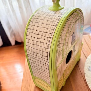 May include: A decorative birdhouse with a cage design. The birdhouse is painted in shades of green and cream, with a black grid pattern and a small bird illustration. The word "maison" is written on the front.